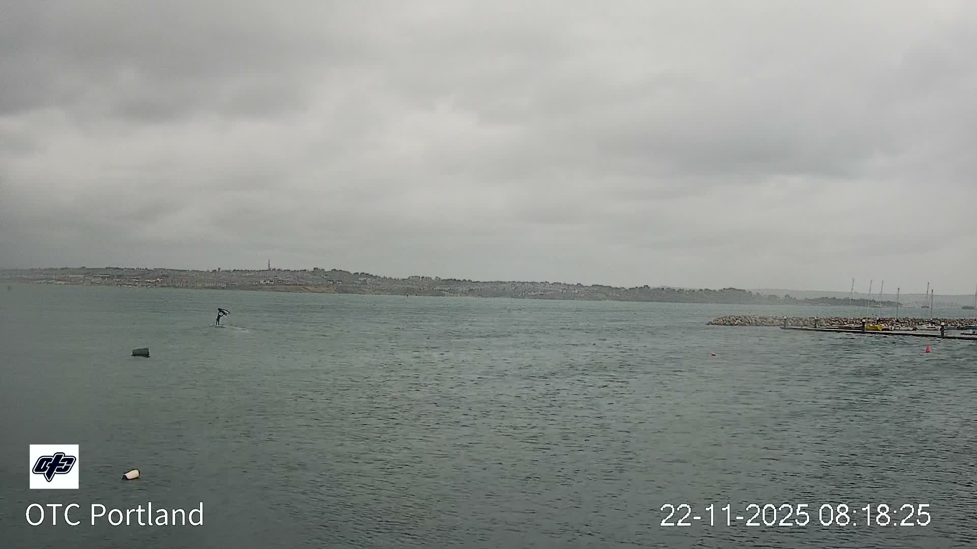 A person is wingfoiling on choppy grey water under an overcast sky, with a distant town and a marina visible in the background.