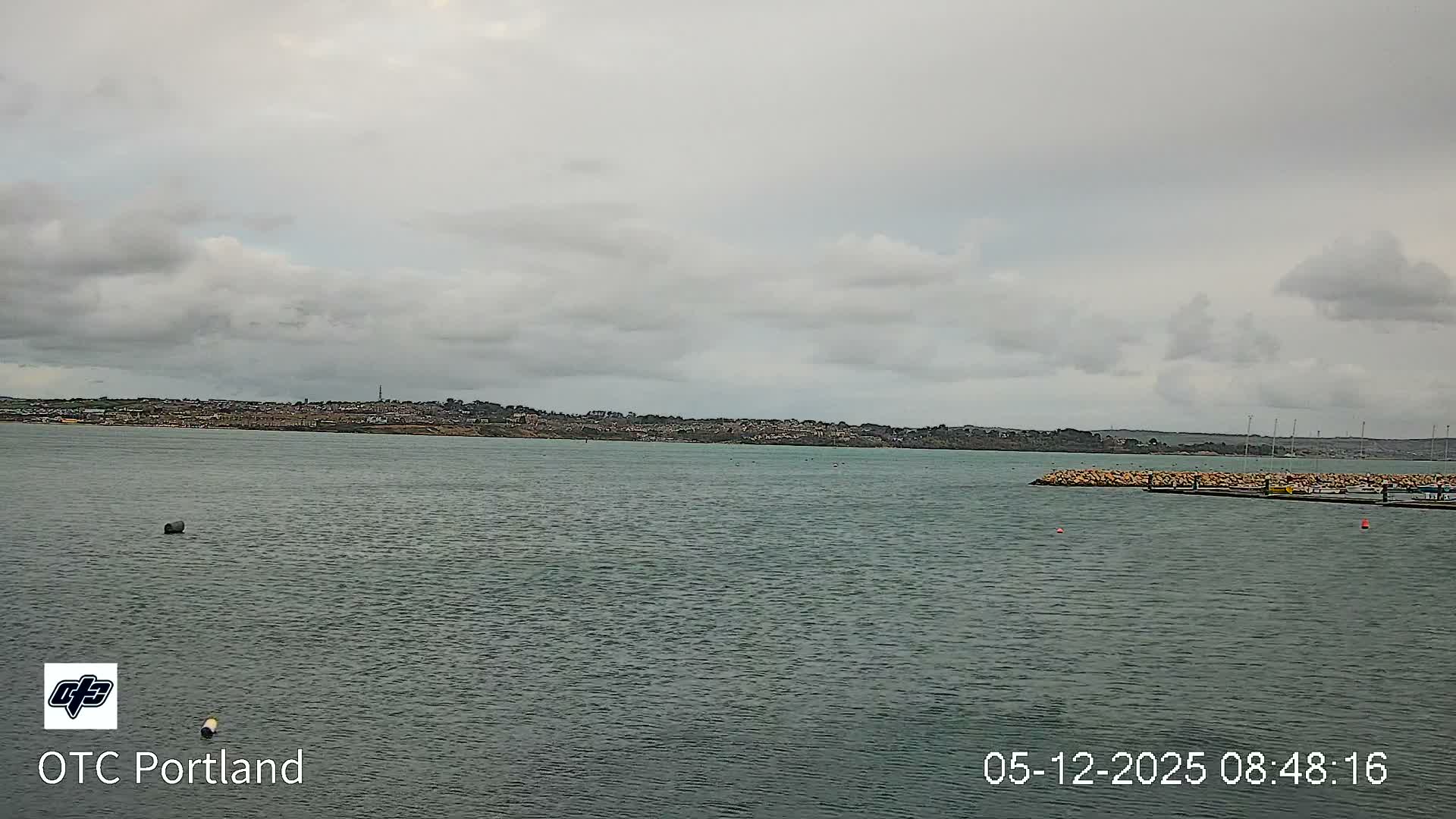 A wide view shows a calm body of water with scattered buoys in the foreground and a rock-protected marina on the right, all set against a distant, built-up coastline under a uniform overcast sky.
