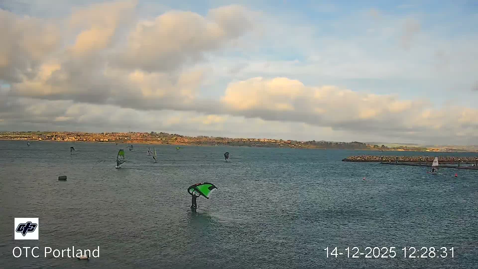 A wide view shows a calm body of water with scattered buoys in the foreground and a rock-protected marina on the right, all set against a distant, built-up coastline under a uniform overcast sky.