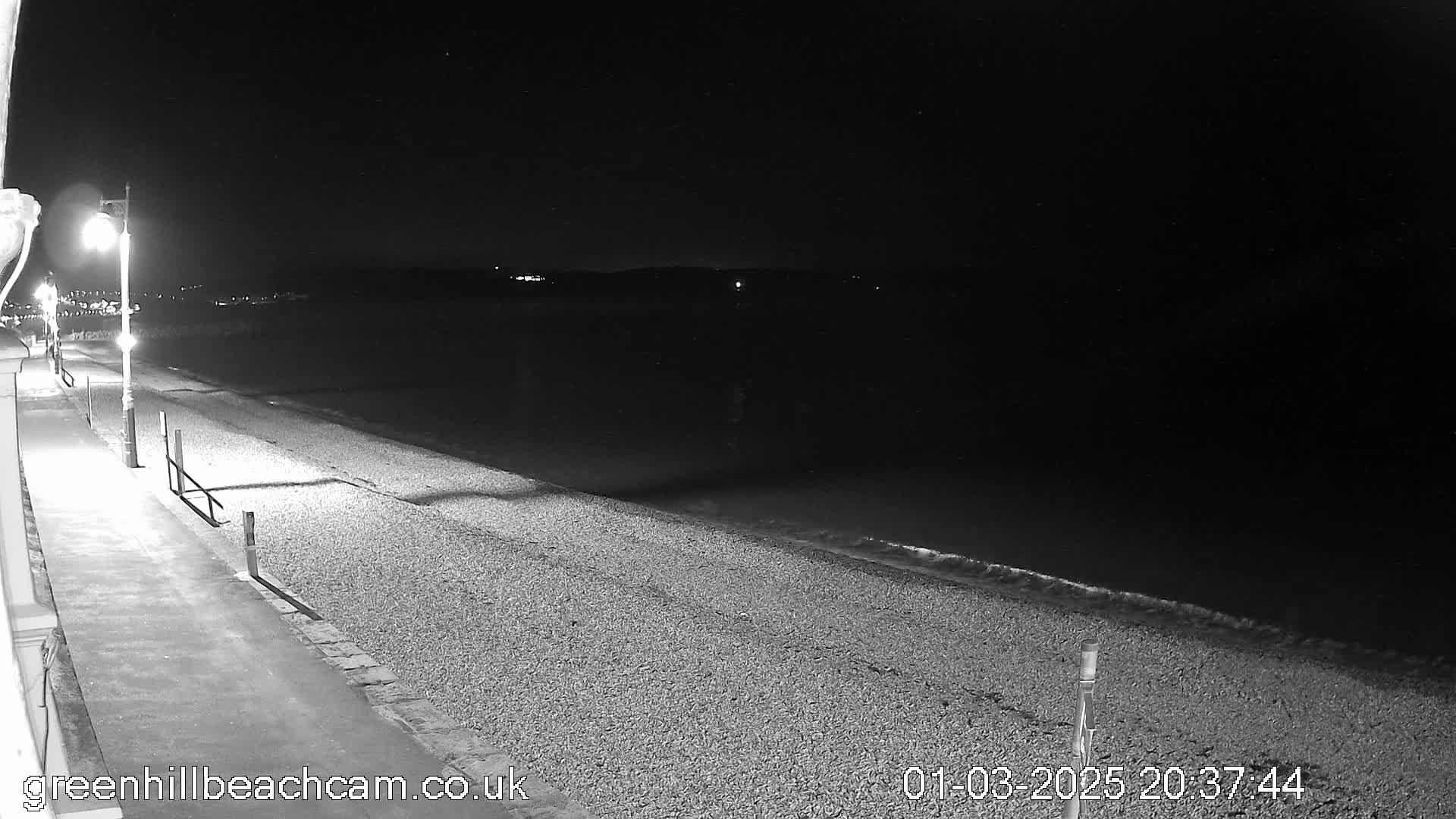 A nighttime view of a pebble beach with gentle waves lapping the shore, illuminated by streetlights along a paved walkway.