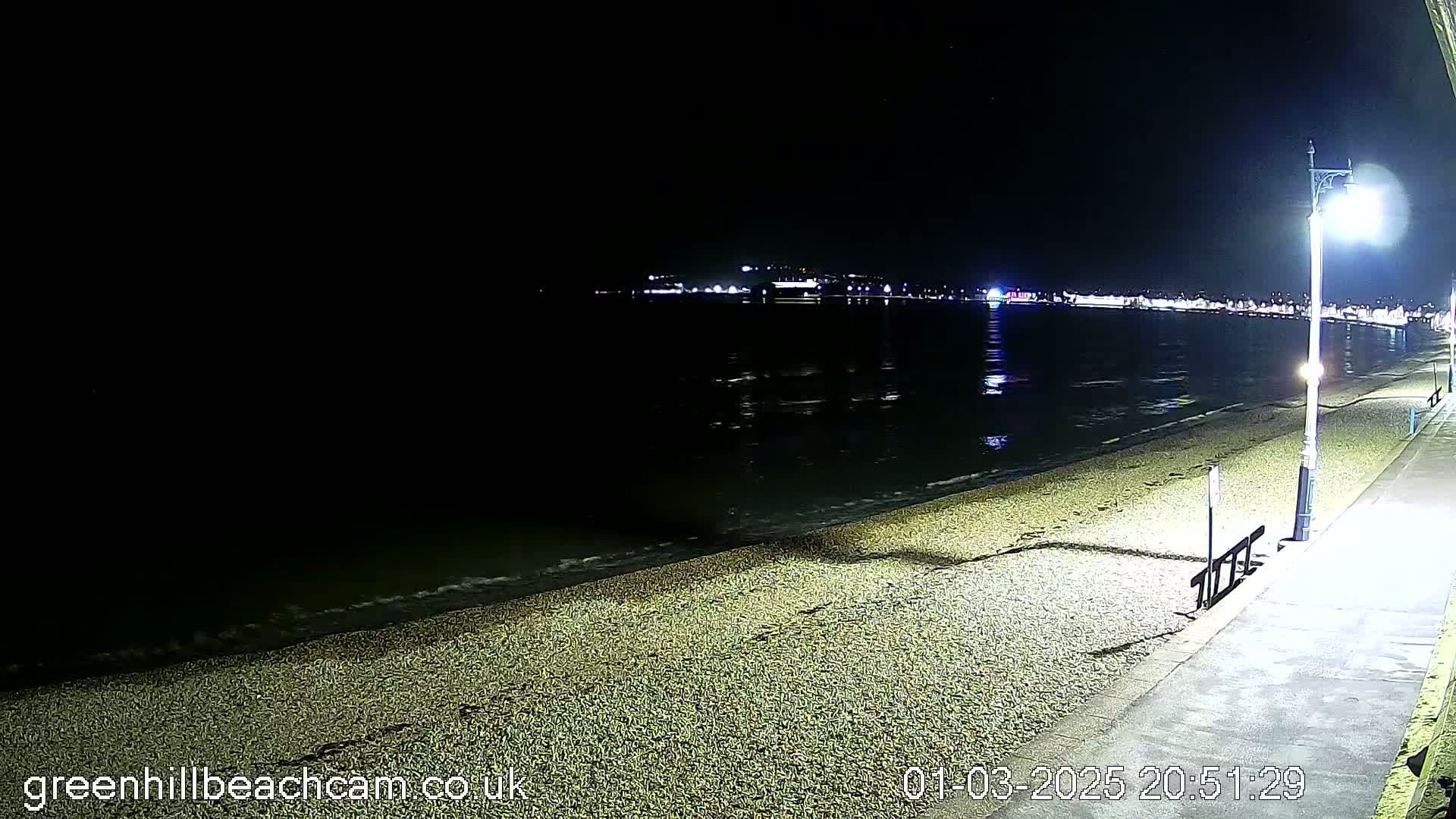 A dark, nighttime beach scene shows a pebble shoreline with gentle waves lapping at the sand under a mostly clear sky, and a paved walkway with a streetlight along the edge, and lights visible across the water in the distance.