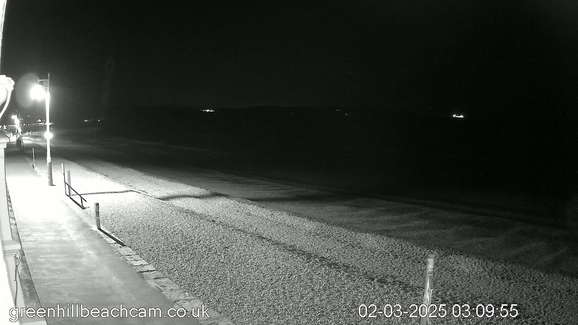 A nighttime, black and white view shows a pebble beach and a paved walkway illuminated by a streetlight, with a dark ocean extending to a distant, faintly lit shoreline.