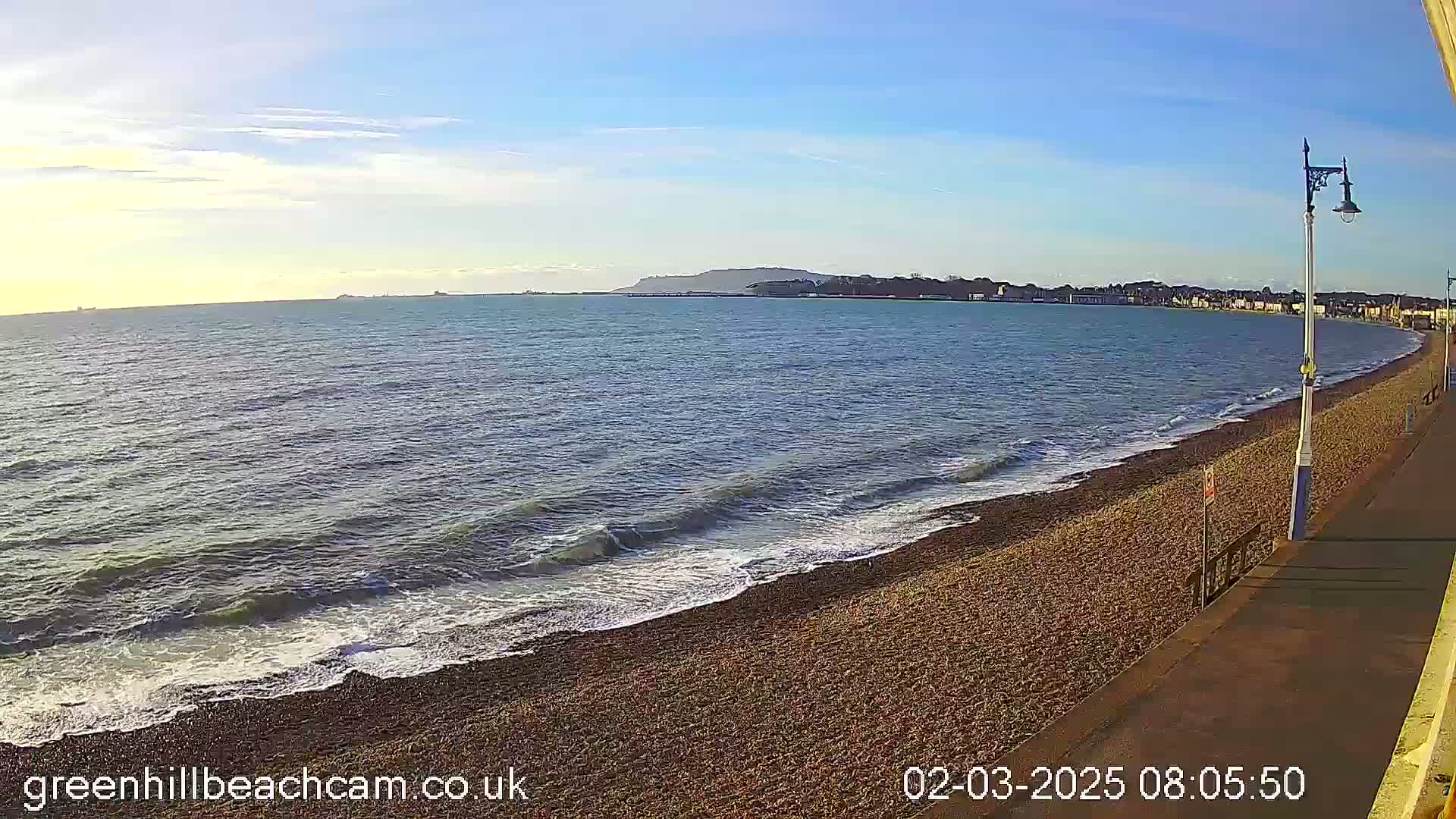 A pebble beach with gentle waves under a mostly clear blue sky stretches to a distant coastline with a hill.