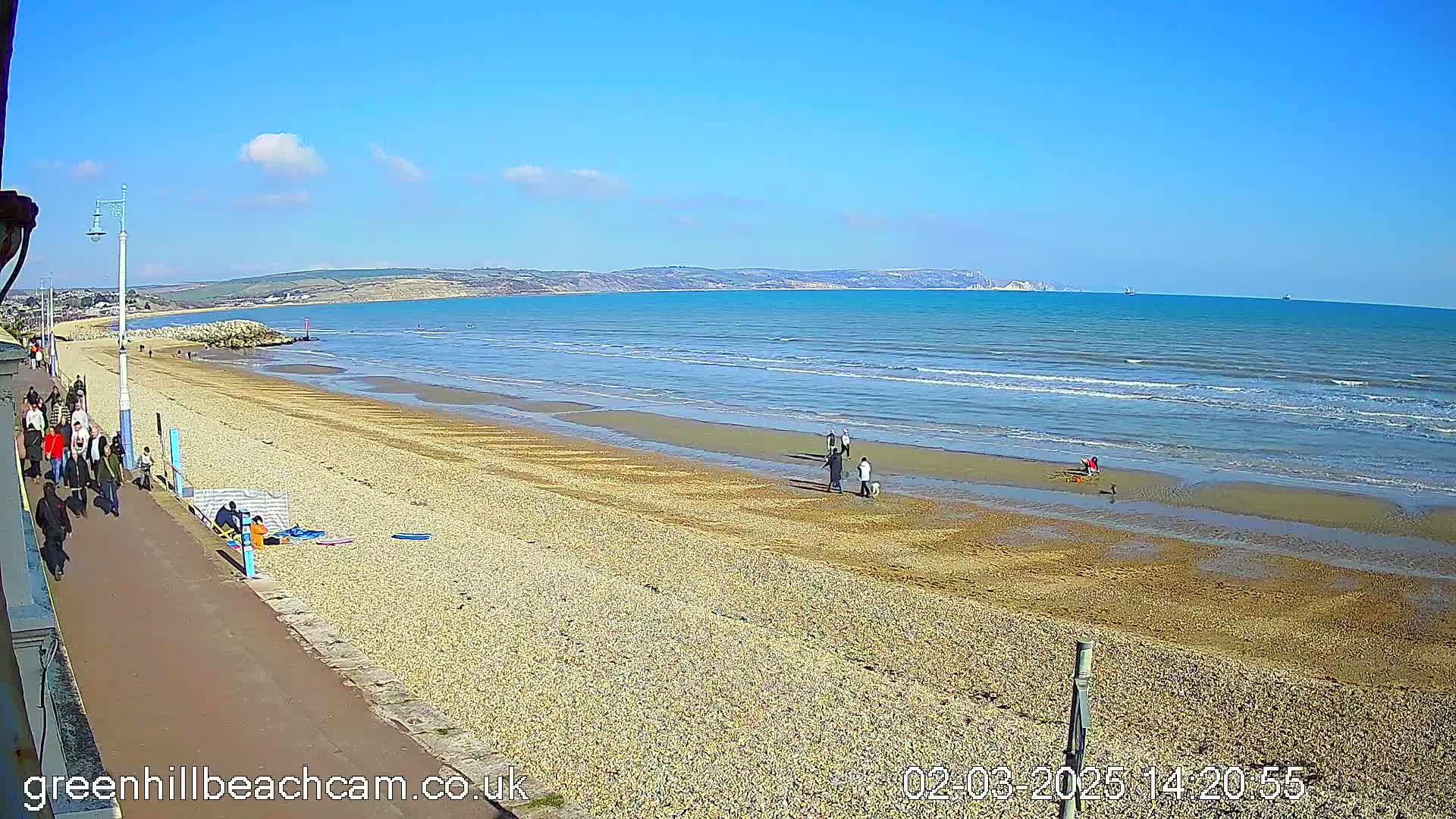 A sunny day at a pebble beach shows people walking along a paved path and strolling on the sand near calm, blue water with distant hills visible on the horizon.