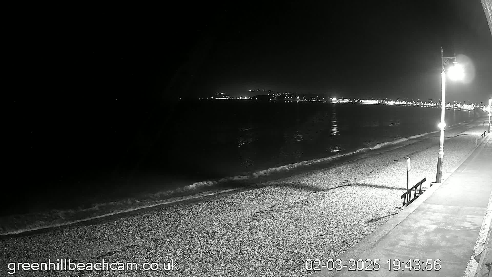A dark, nighttime beach scene shows gently lapping waves on a pebble shore, illuminated by a streetlight, with distant city lights visible across the water.