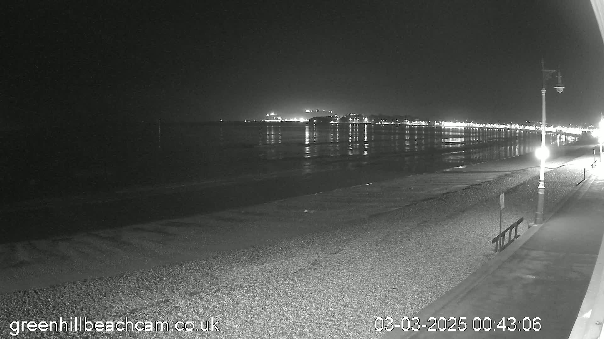 A nighttime view of a calm, dark beach with lights reflecting in the water and a town in the distance.