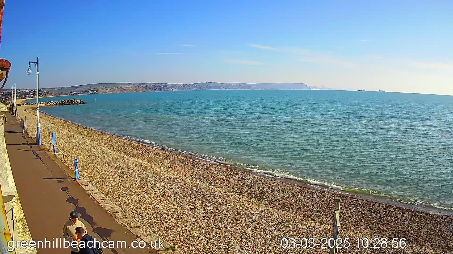 A pebble beach stretches along a calm, turquoise sea under a clear blue sky, with two people walking along a paved path beside it.