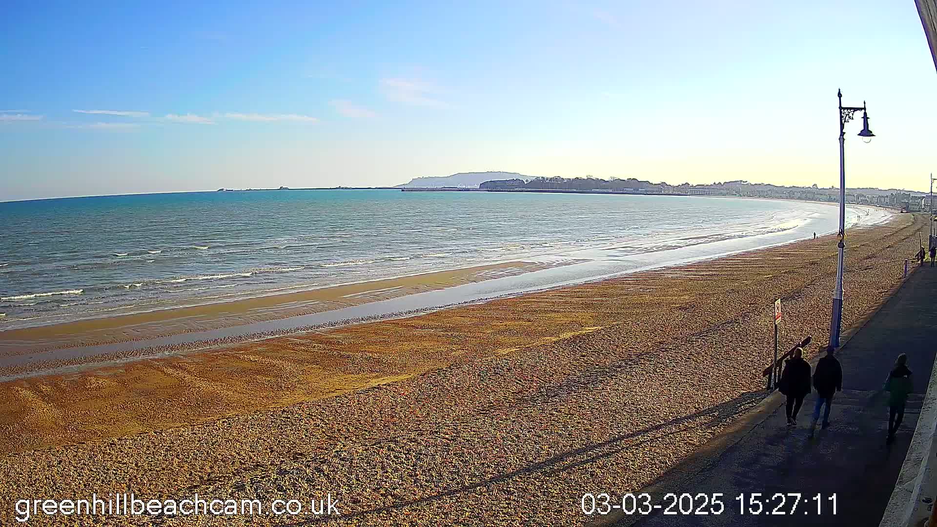 A long, sandy beach stretches along a calm sea under a clear blue sky, with a few people walking along a paved path near the shoreline.