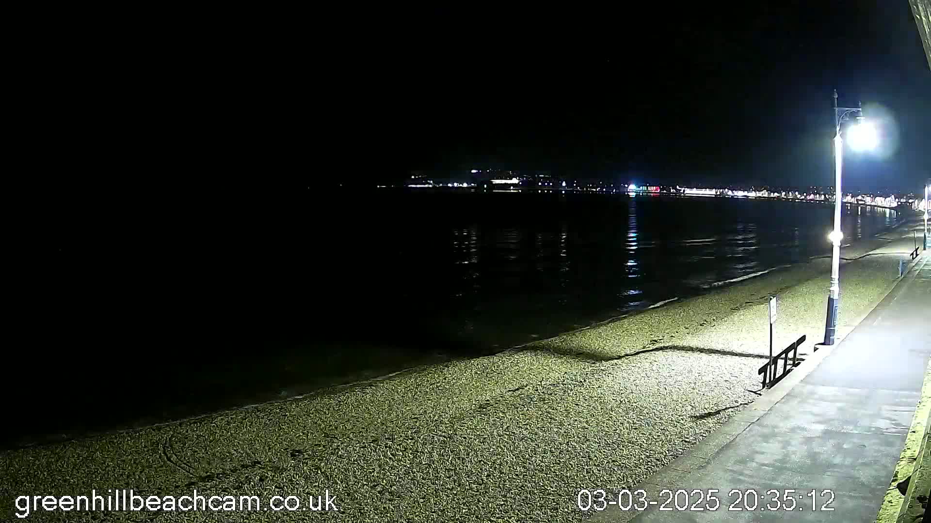 A dark, pebble beach at night is illuminated by a streetlight, with a calm body of water and distant lights across the bay.