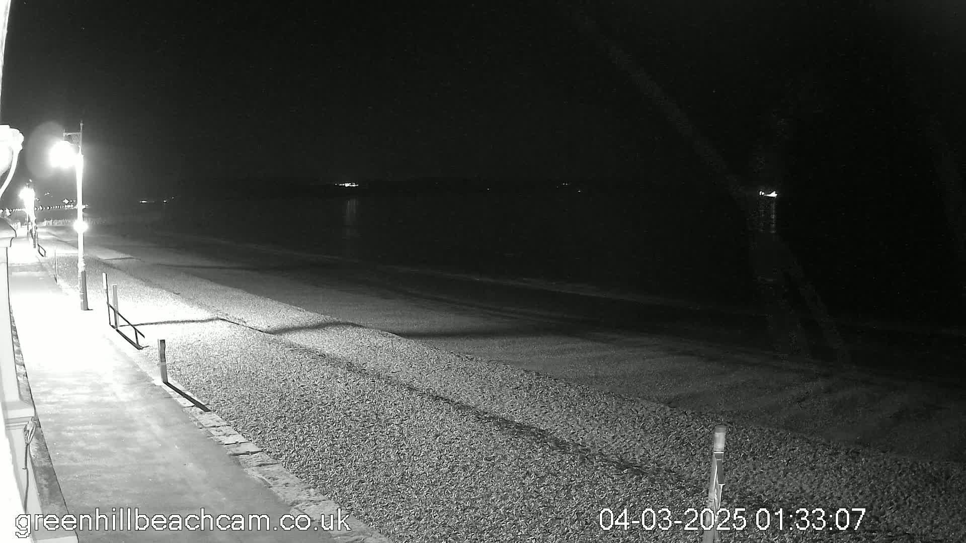A nighttime, black-and-white view shows a pebble beach and a paved walkway next to it, illuminated by a streetlight, with calm water visible in the distance.