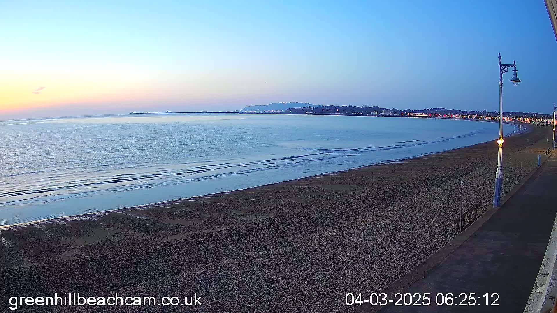 A calm, twilight ocean meets a dark, pebbled beach and a paved walkway lined with lampposts.