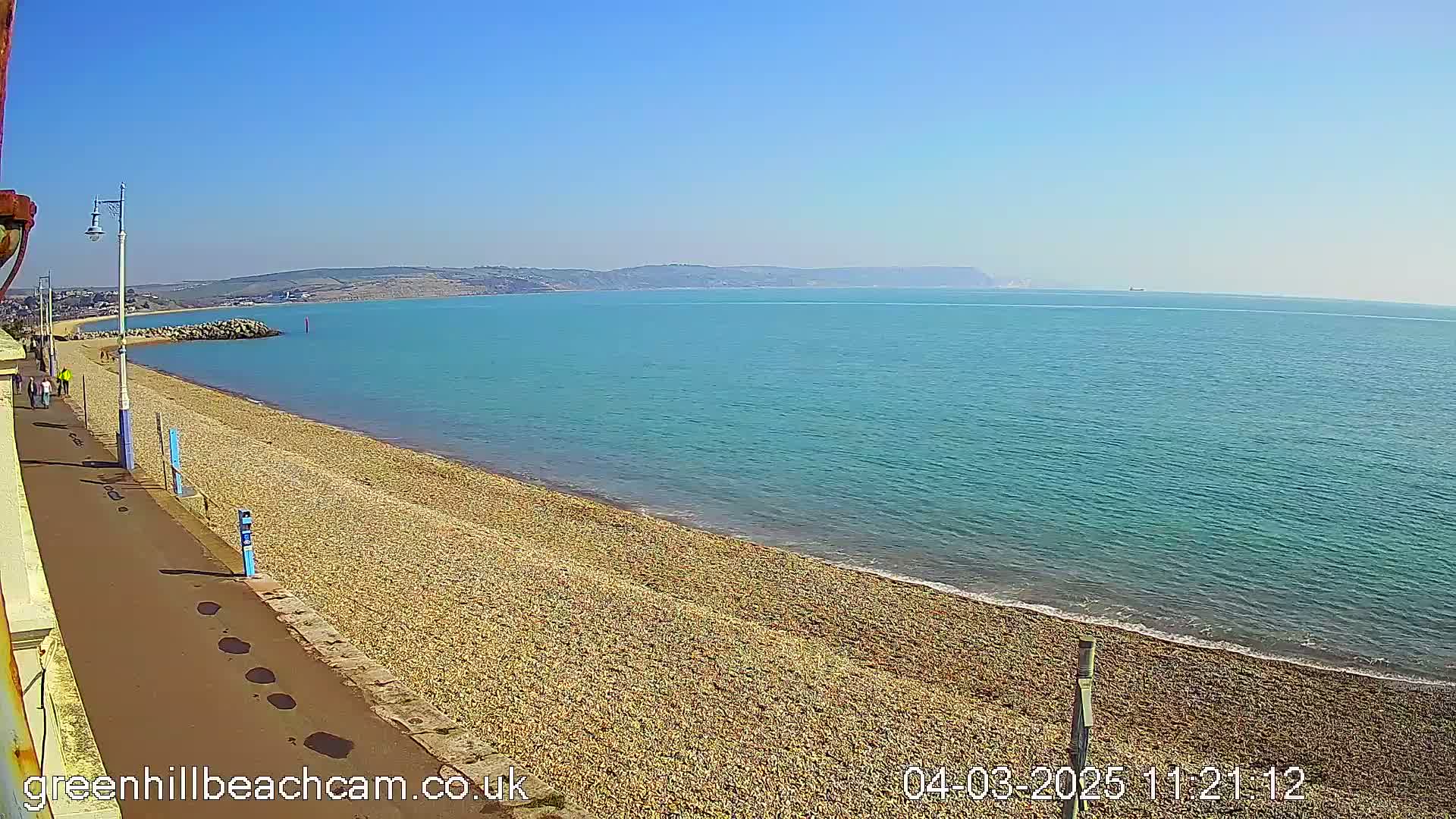 A pebble beach stretches along a calm, teal sea under a clear blue sky, with a paved walkway and distant hills visible.
