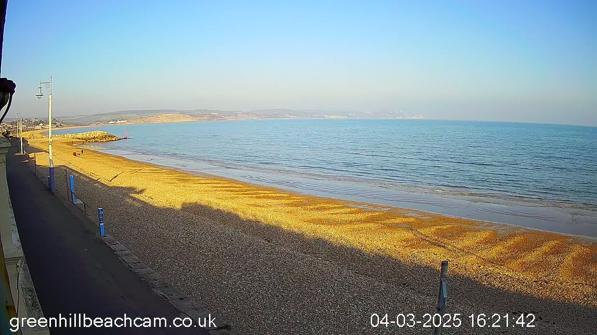 A mostly sunny day at a pebble beach shows a calm sea with a long, sandy shoreline and a paved walkway next to it.