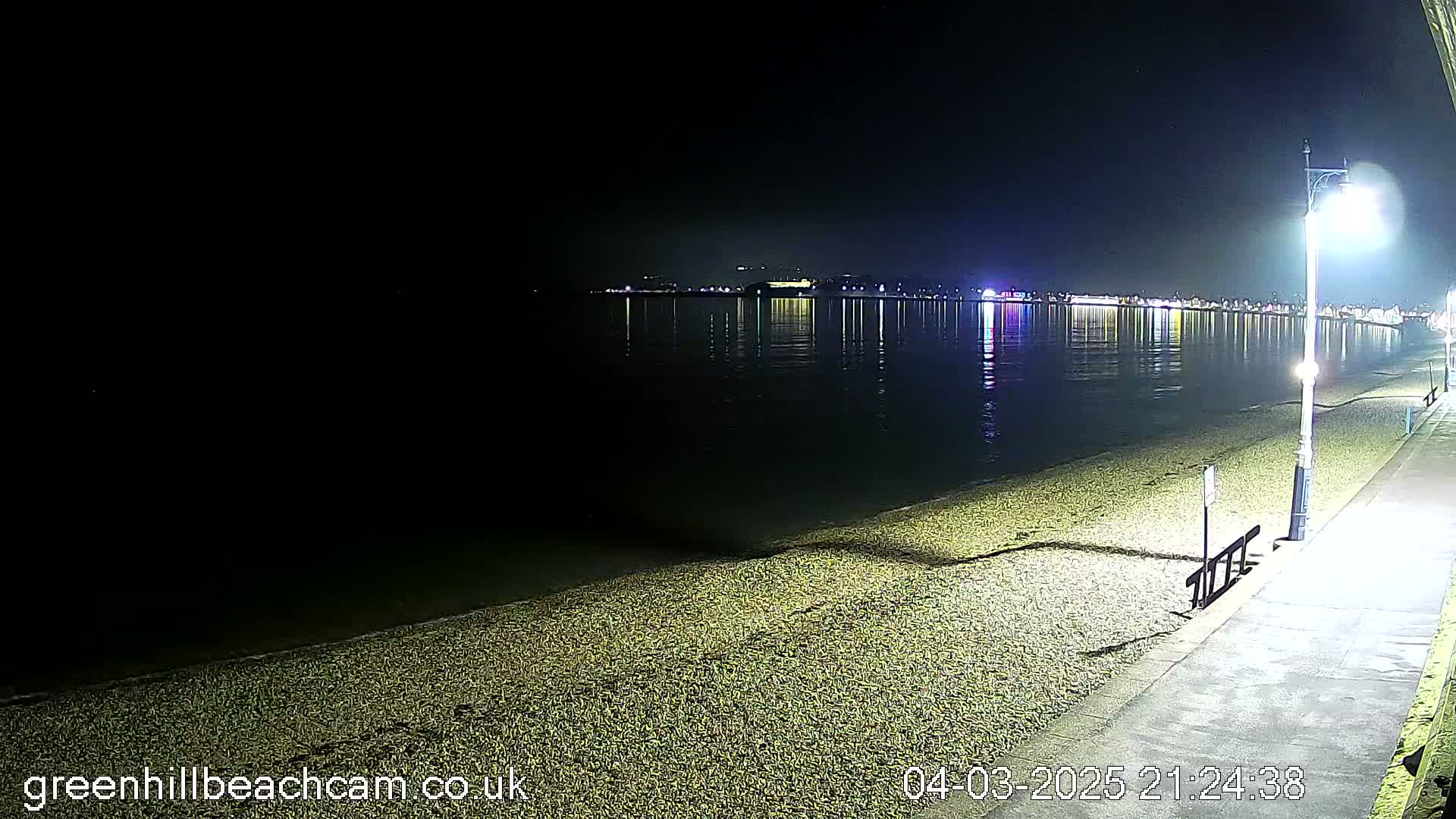 A nighttime view of a pebble beach and calm water reflecting city lights under a clear, dark sky.