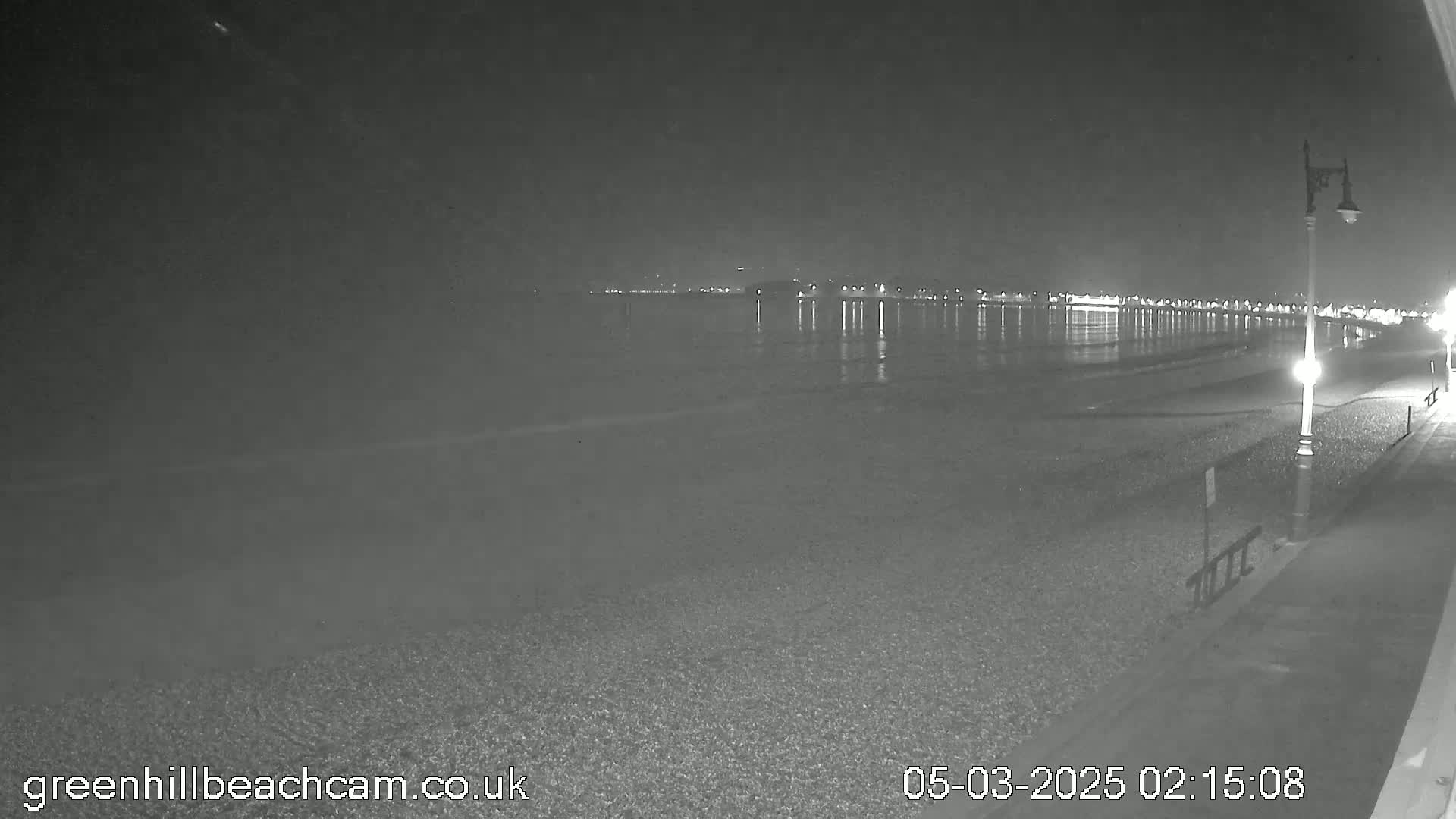 A nighttime view of a calm, dark ocean reflecting lights from a distant town on a pebble beach, illuminated by a single lamppost.