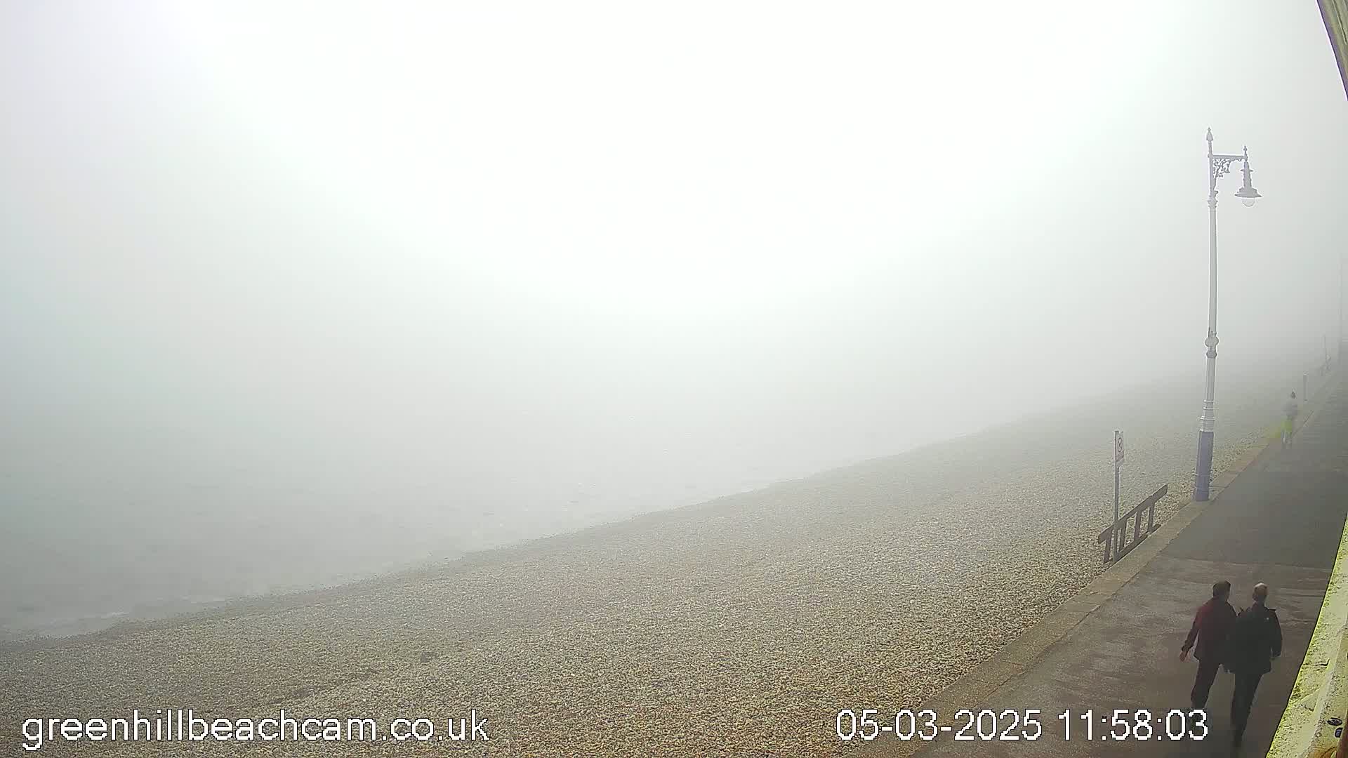 Two people walk along a foggy beachside promenade next to a pebble beach and the sea, under a lamppost.