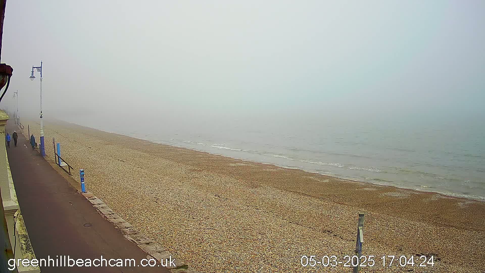 A foggy day at a pebble beach shows a paved walkway with a few people walking, alongside a calm sea.