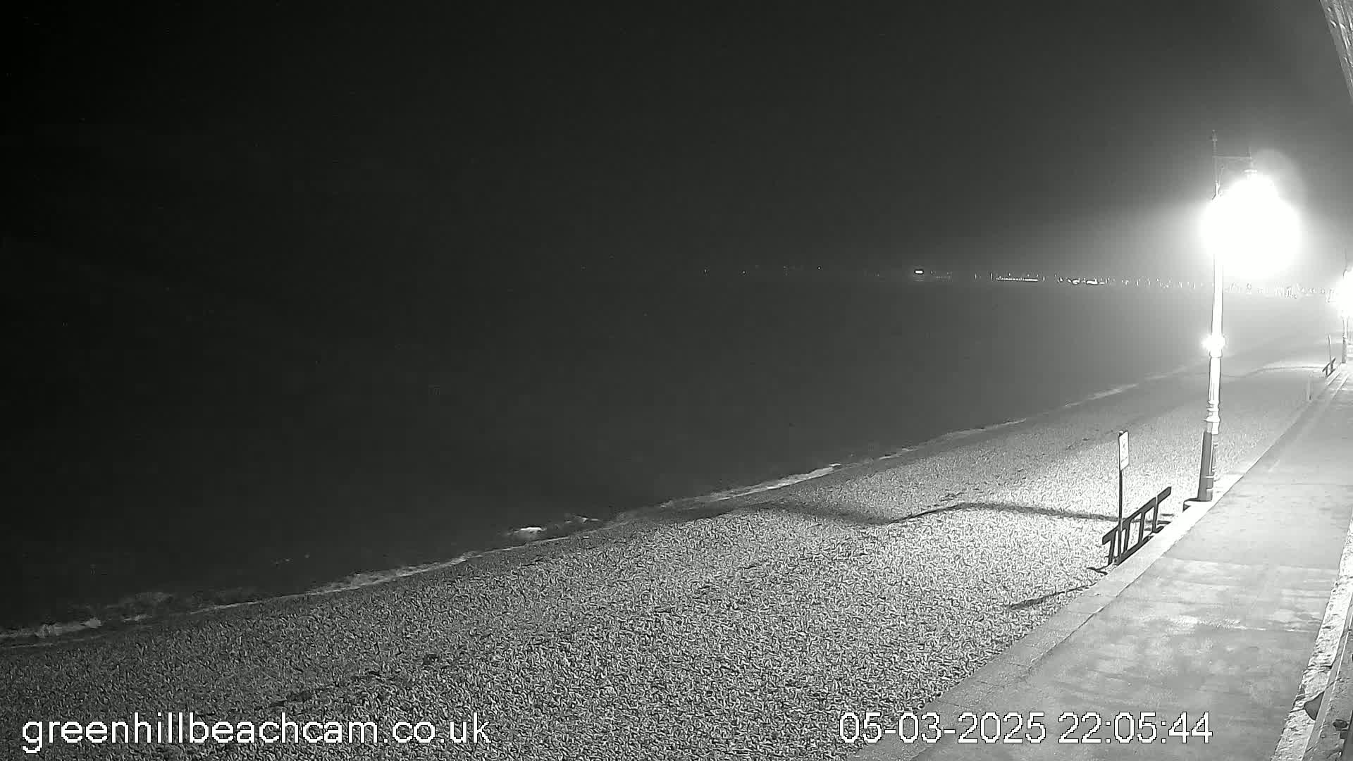 A dark, nighttime beach scene shows a pebble shoreline gently lapping at the water's edge under a single illuminated lamppost.