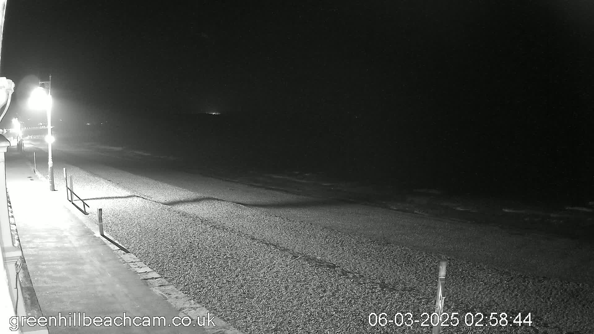 A nighttime, black and white view of a pebble beach and paved walkway illuminated by a streetlight, with calm ocean waves visible in the background.