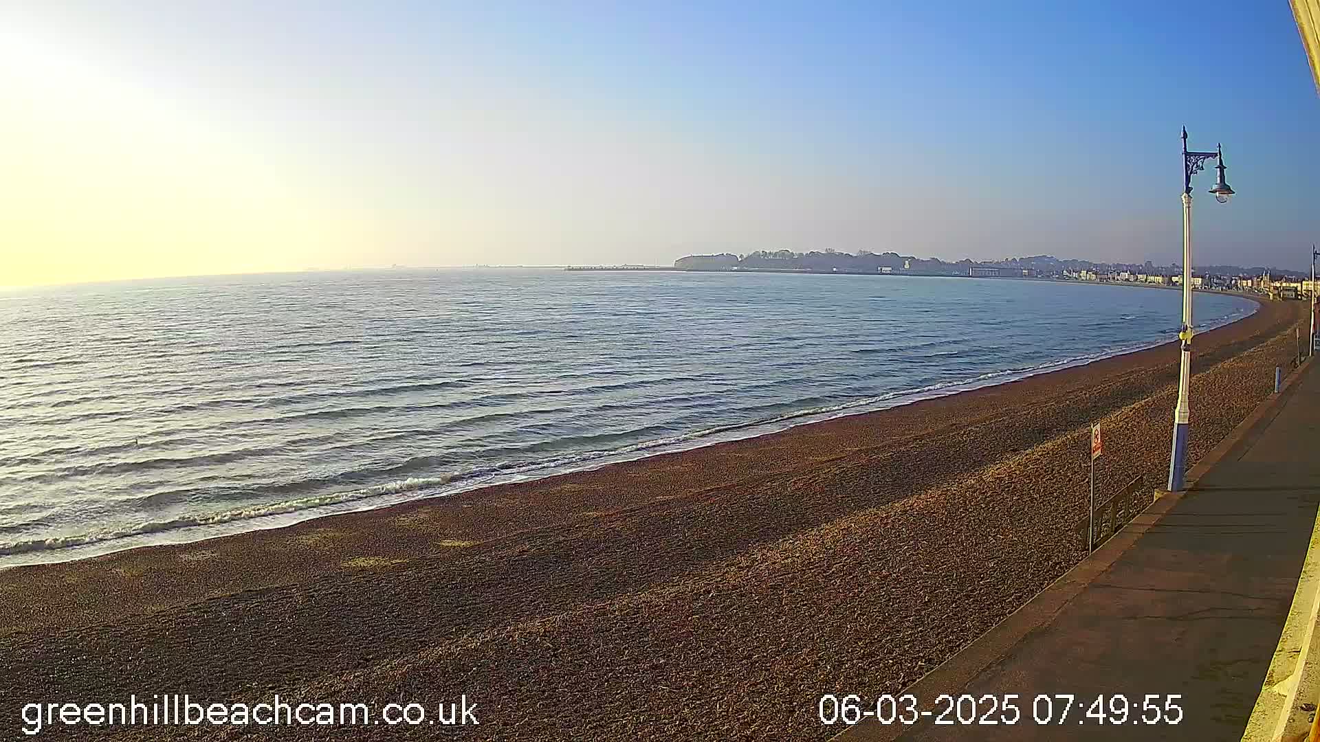 A calm, clear morning shows a gently sloping pebble beach next to a paved walkway, with a calm sea stretching to a distant shoreline under a clear blue sky.
