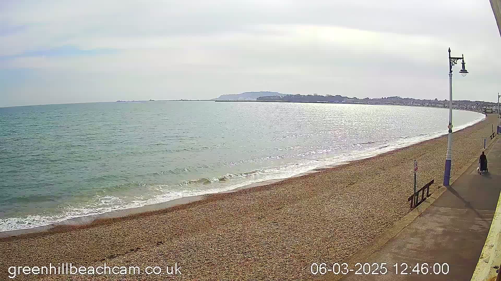 A gently sloping pebble beach meets a calm sea under a mostly cloudy sky, with a person and a dog walking along a paved walkway beside the beach.