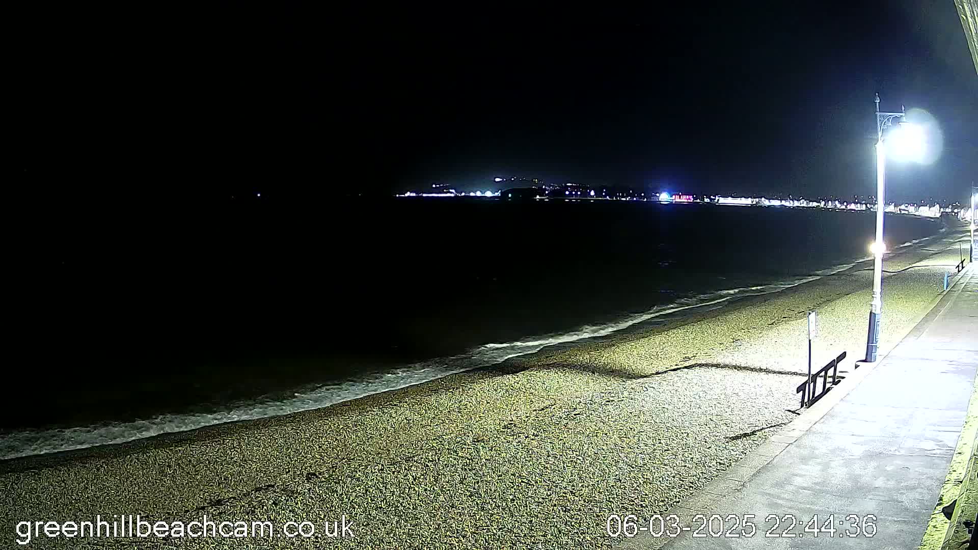 A dark, nighttime beach scene shows gently lapping waves on a pebbled shore, illuminated by a streetlight on a paved walkway next to the beach, with city lights visible in the distance.