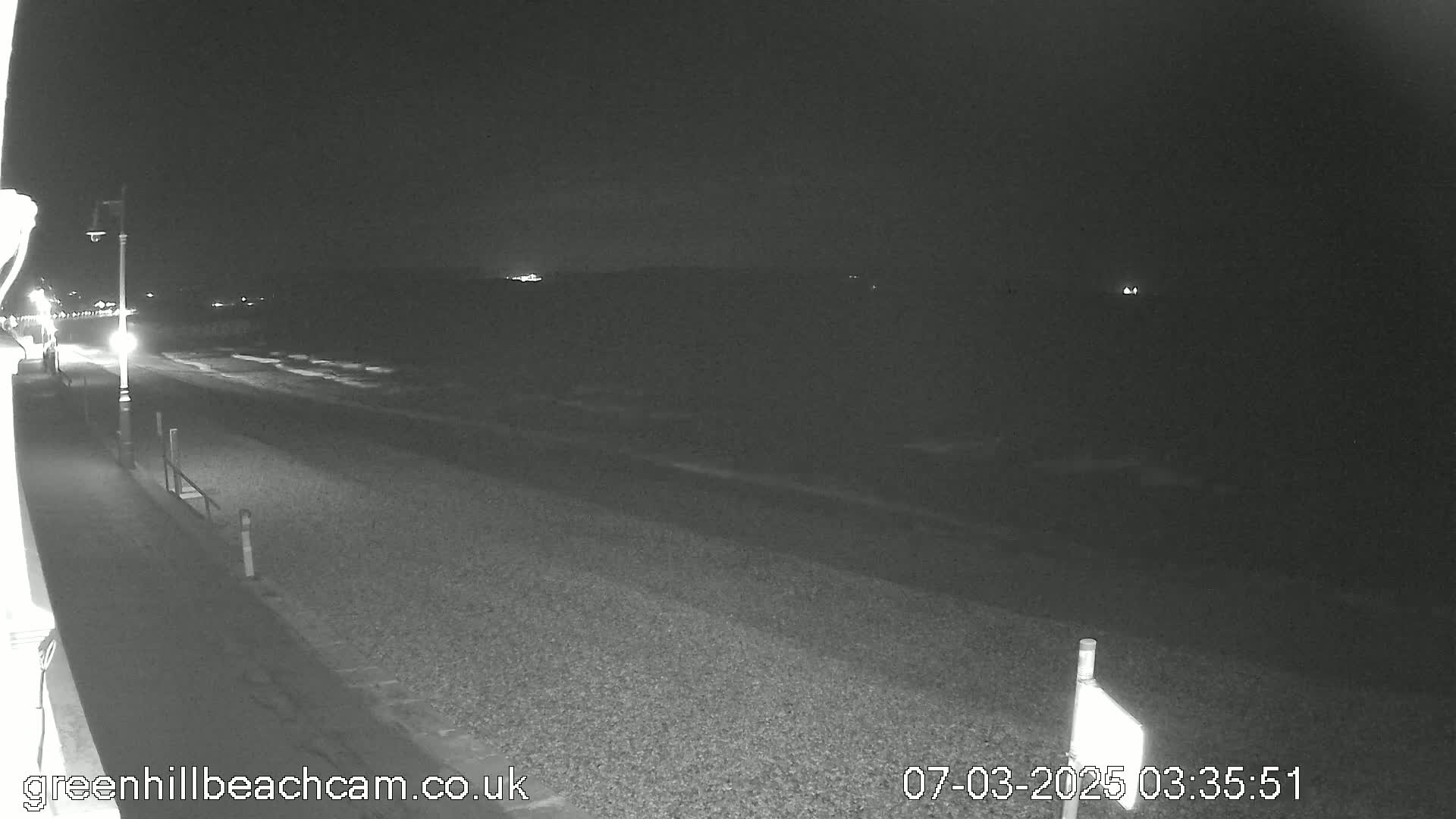 A dark, nighttime view of a pebble beach and a paved walkway with a single lamppost, under a clear sky.