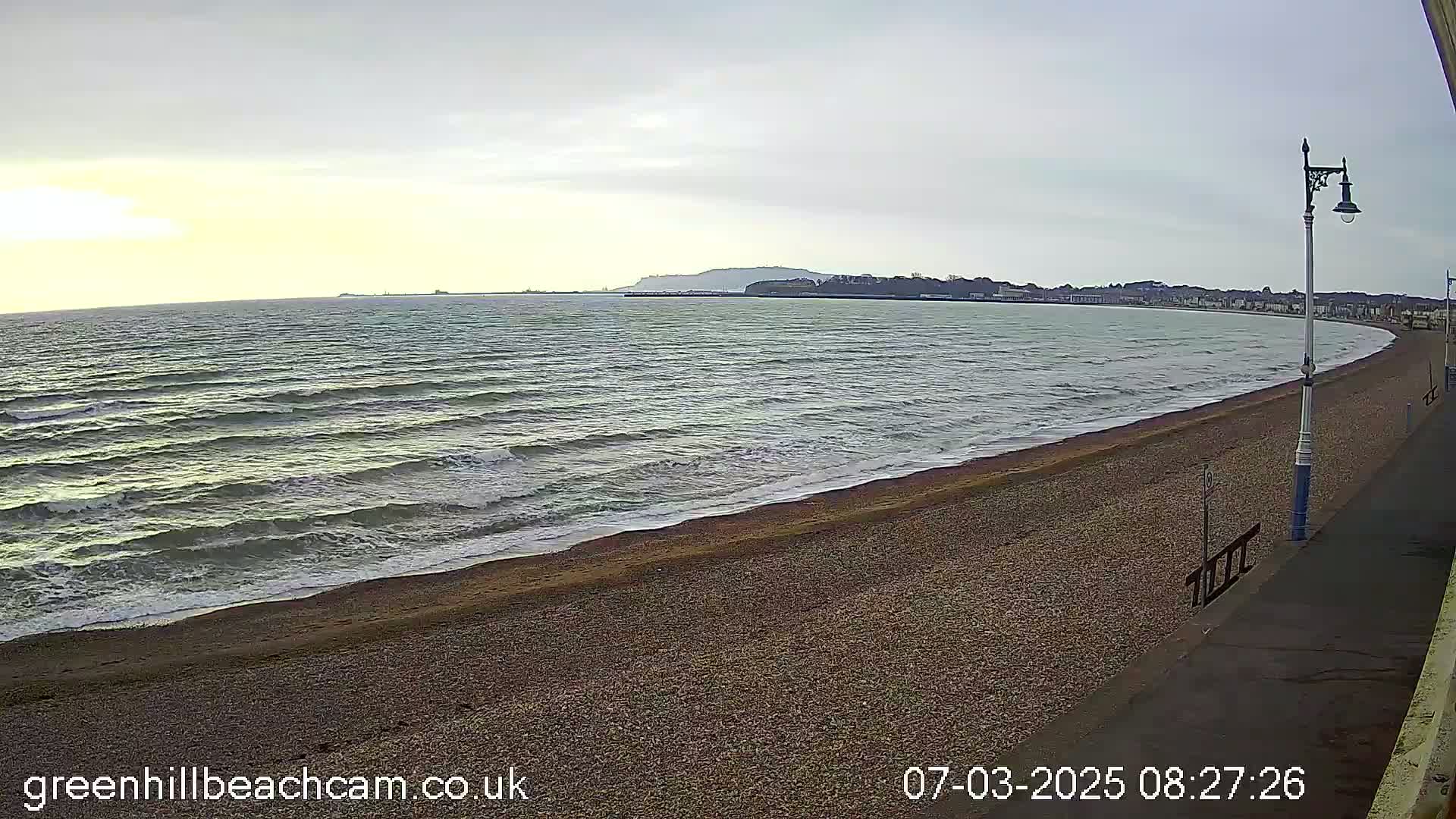 A pebble beach curves gently around a calm, gray sea under an overcast sky.