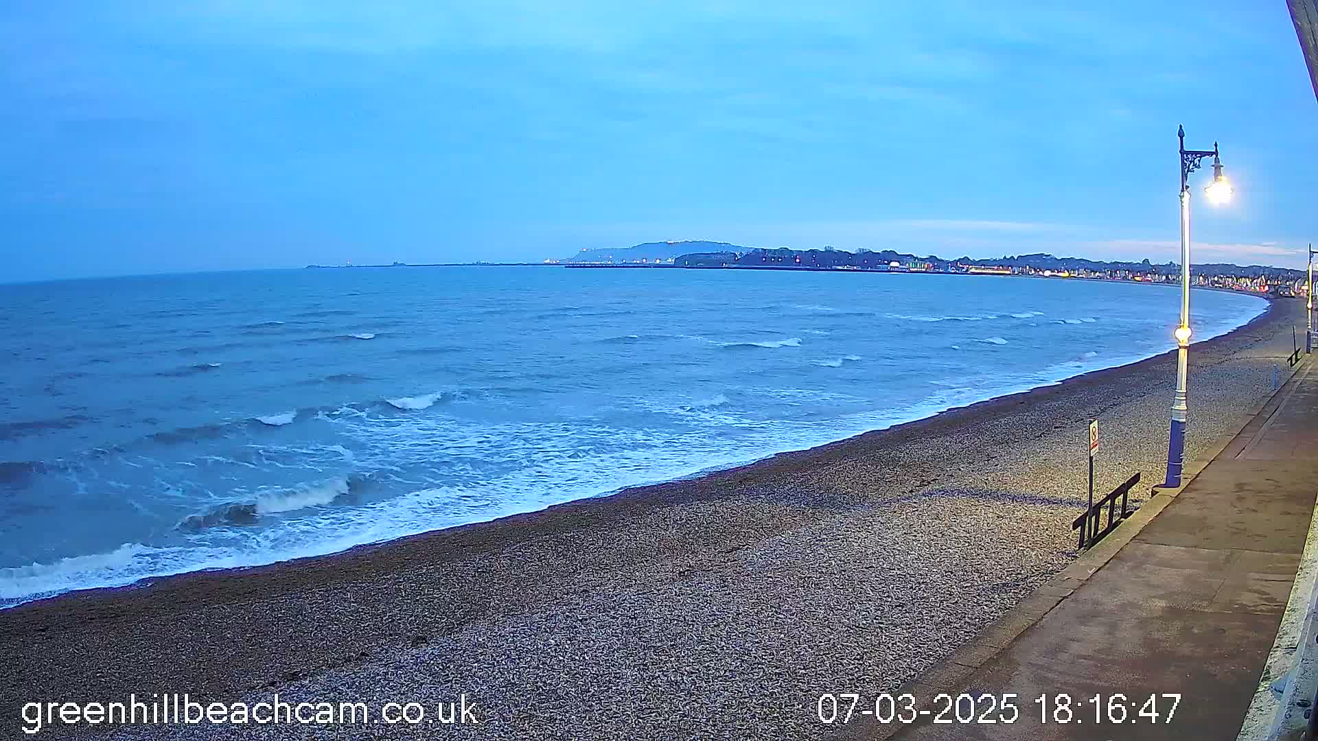 A pebble beach at dusk with small waves rolling onto the shore, a paved walkway nearby, and a town visible in the distance.