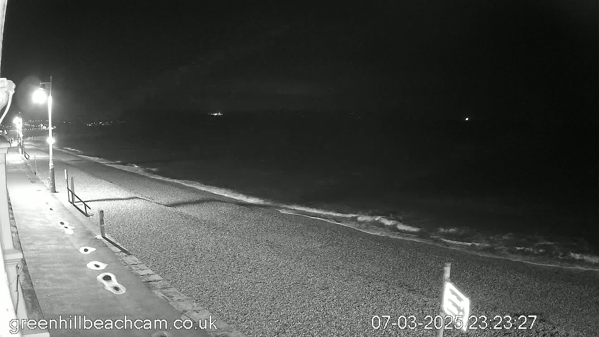 A dark, pebble beach at night with gentle waves lapping the shore, lit by streetlights along a paved walkway.