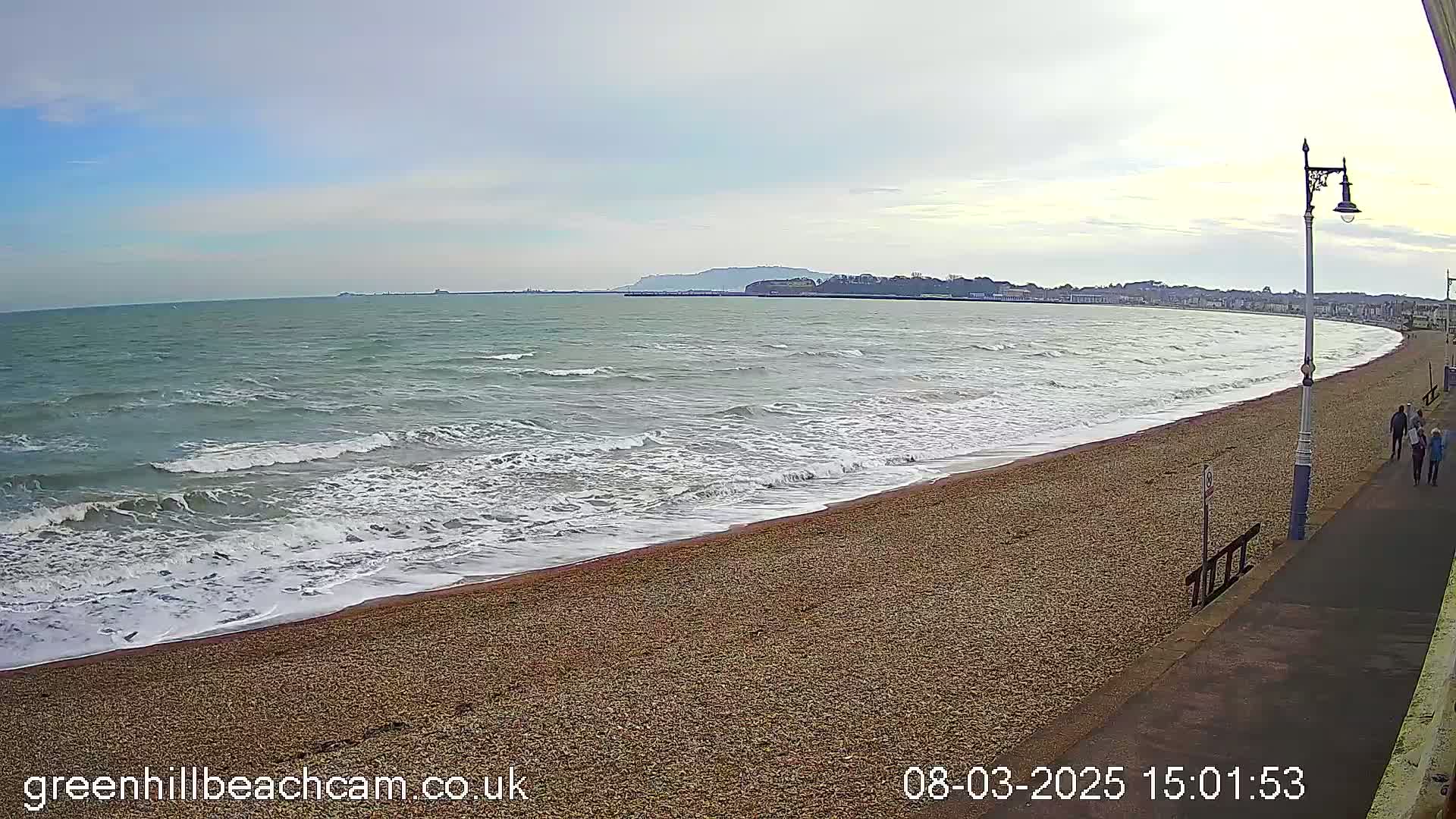 A pebble beach curves gently to the right, with small waves breaking on the shore under an overcast sky.