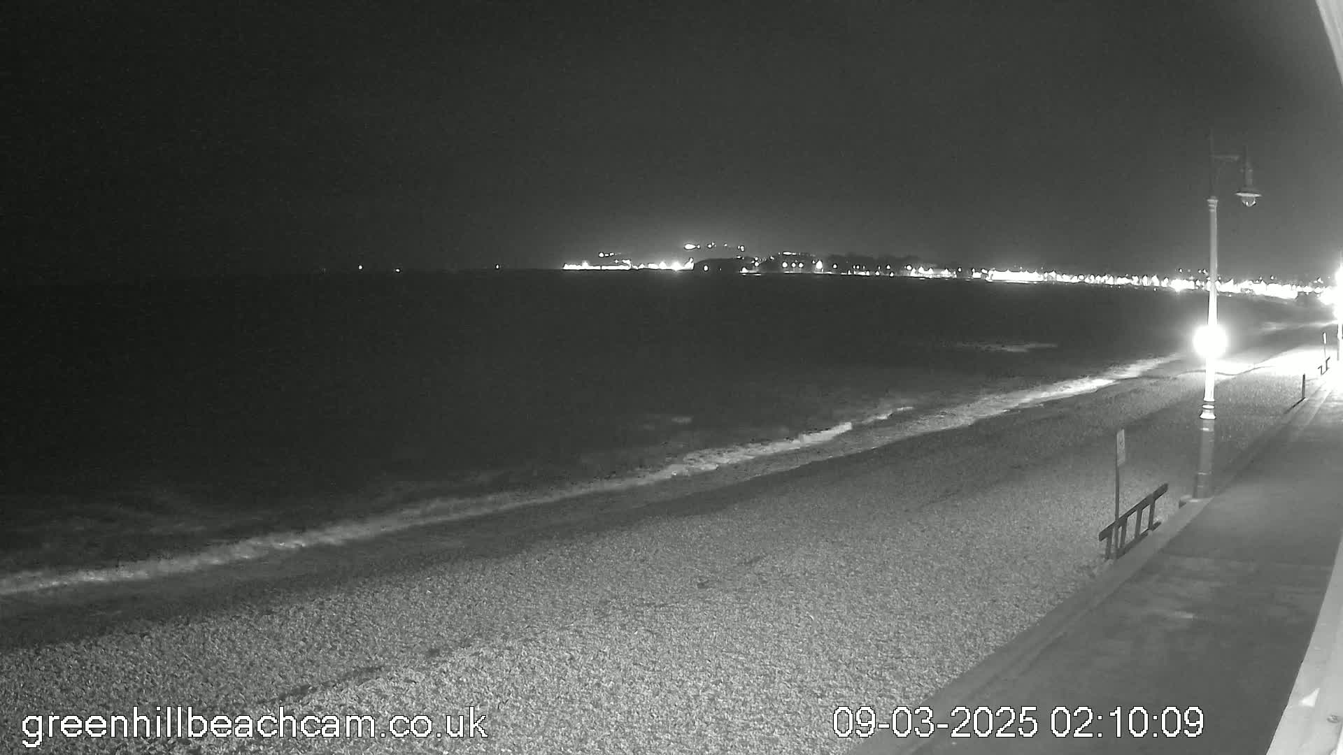 A dark, pebble beach at night with gentle waves lapping the shore, under a clear sky, is lit by streetlights and the distant glow of a town.