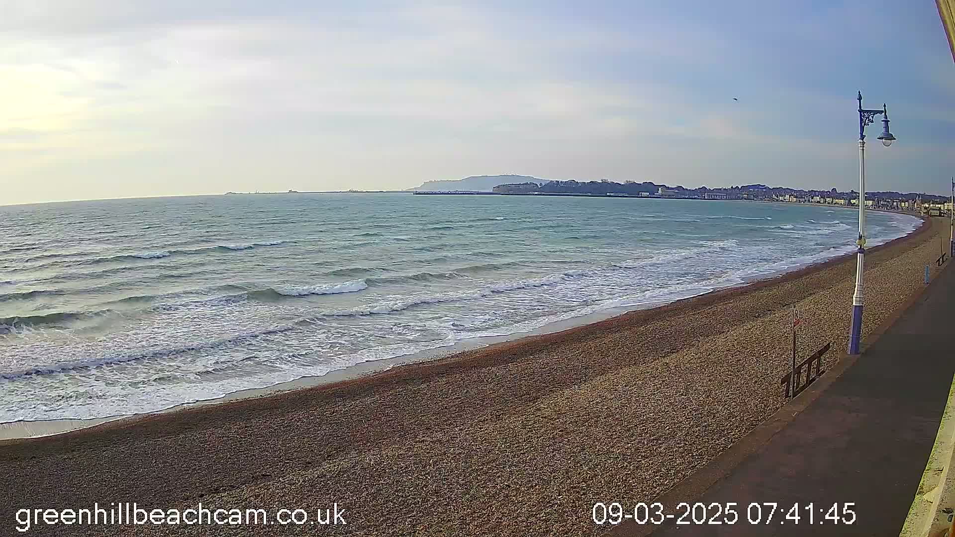 A pebble beach with gentle waves rolling onto the shore under a partly cloudy sky, with a town visible in the distance.
