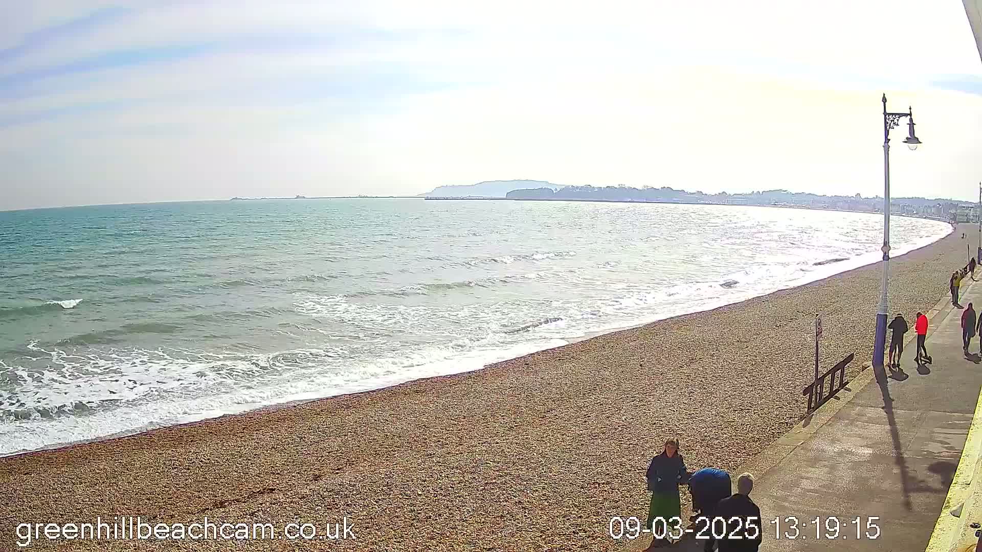 A pebbled beach with gentle waves under a sunny sky features several people walking along a paved walkway next to the water.