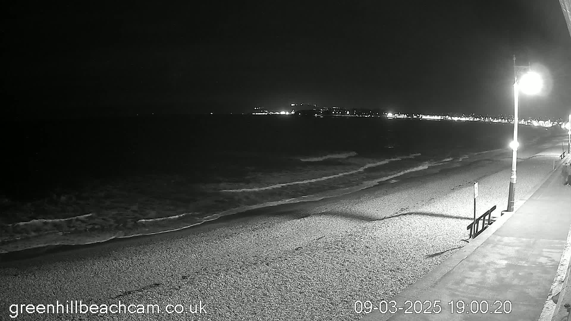 A dark, nighttime beach scene shows gentle waves lapping a pebble shore under a few streetlights, with a town faintly visible in the distance.