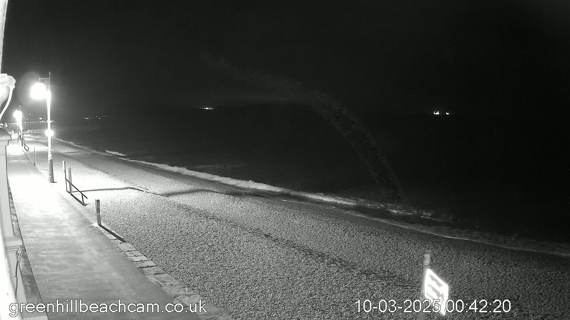 A nighttime view of a pebble beach with gentle waves lapping the shore, illuminated by streetlights along a paved walkway.