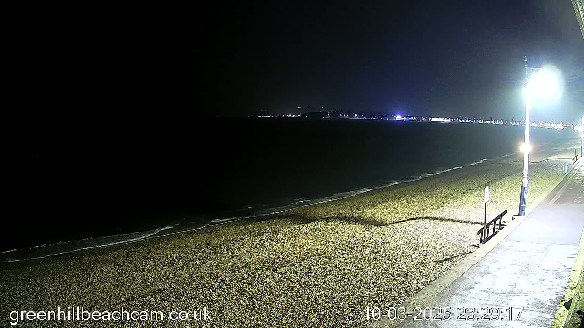 A dark, nighttime beach scene shows gently lapping waves on a pebble shore, illuminated by a streetlight on a paved walkway.