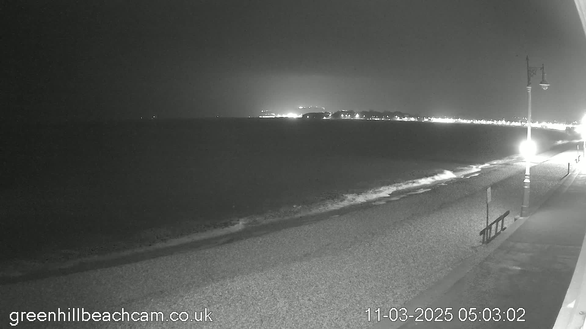 A dark, nighttime view of a pebble beach and calm ocean, with a town faintly lit in the distance.