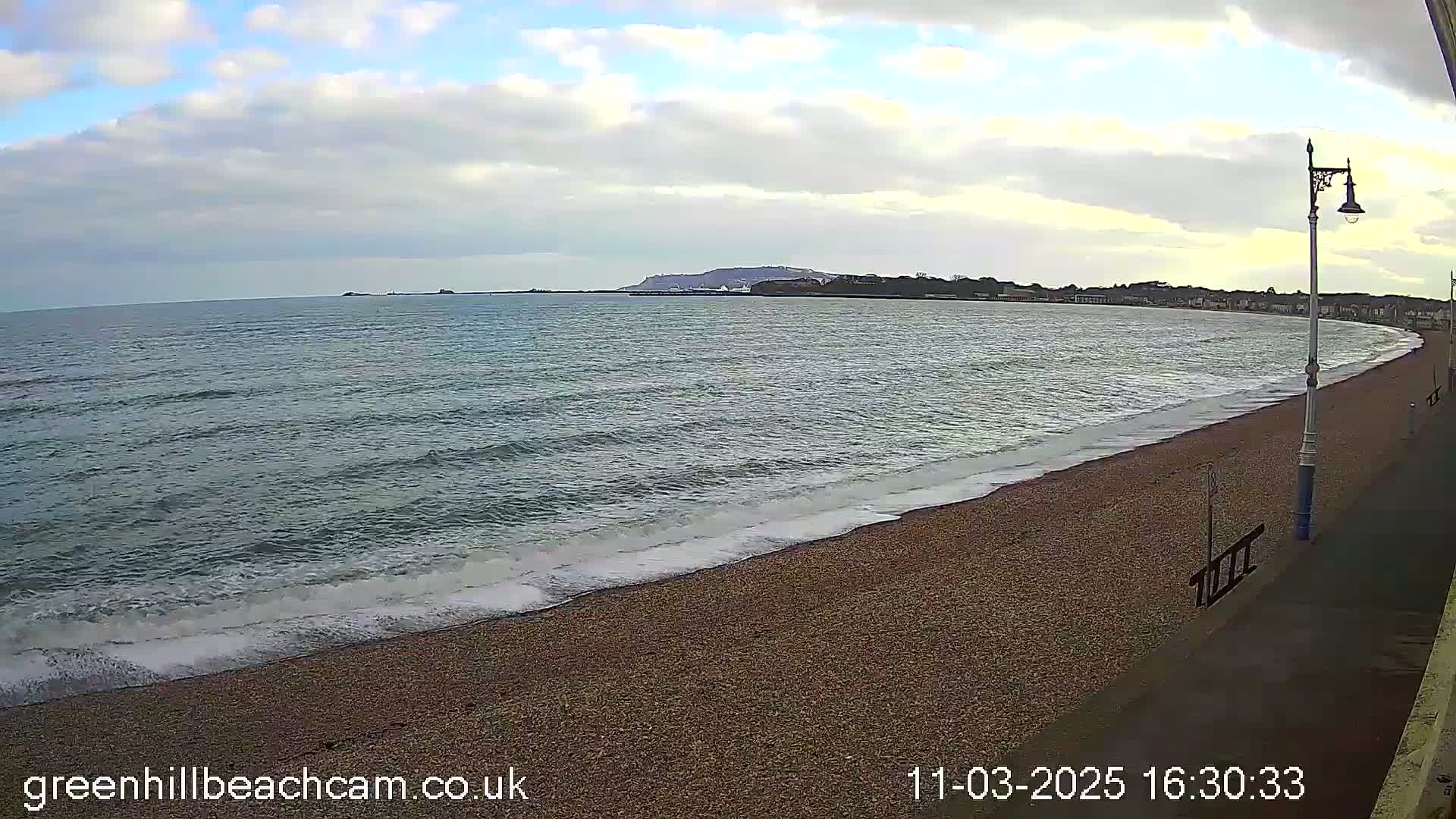 A pebble beach with gentle waves rolls onto the shore under a partly cloudy sky, with a town visible in the distance.