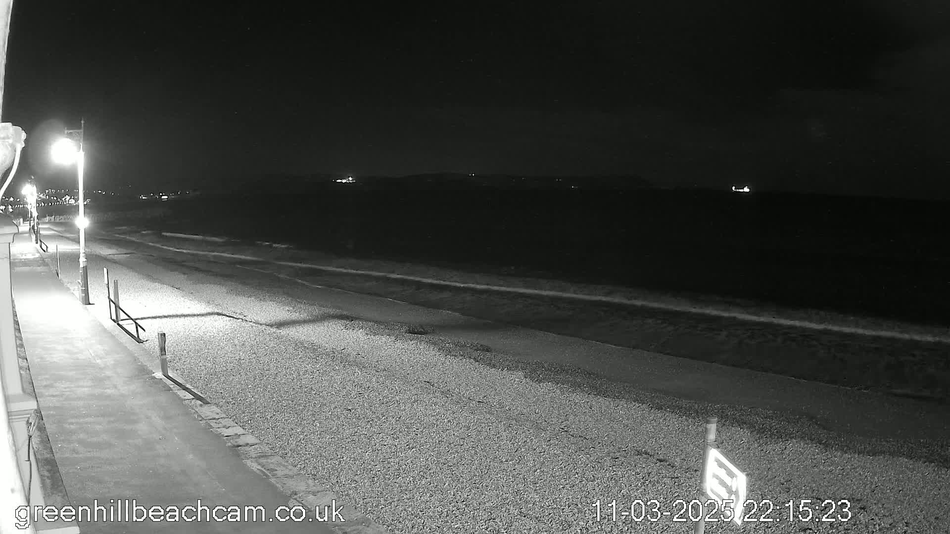 A pebble beach at night with gently lapping waves under a dark sky, lit by a lamppost along a paved walkway.