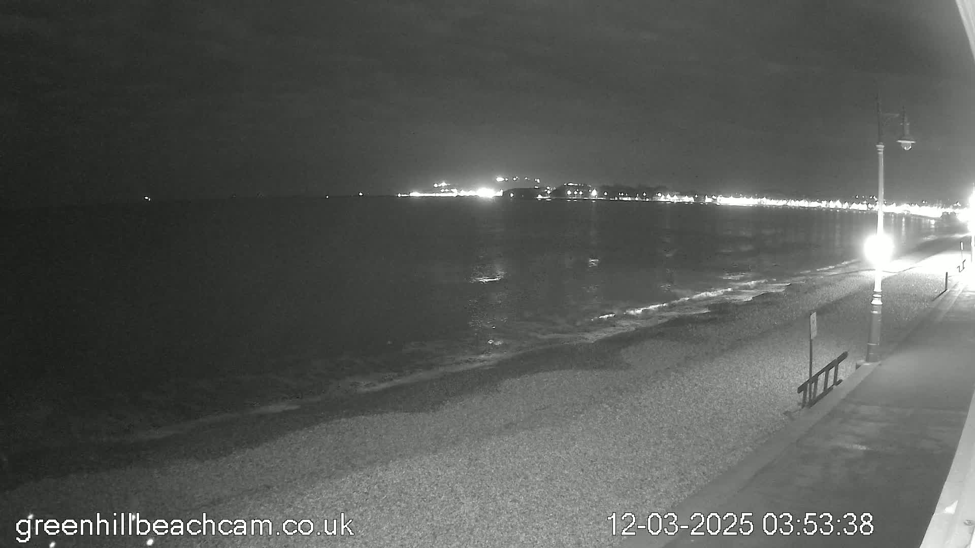 A nighttime view of a pebble beach with calm water and distant city lights under a dark sky.