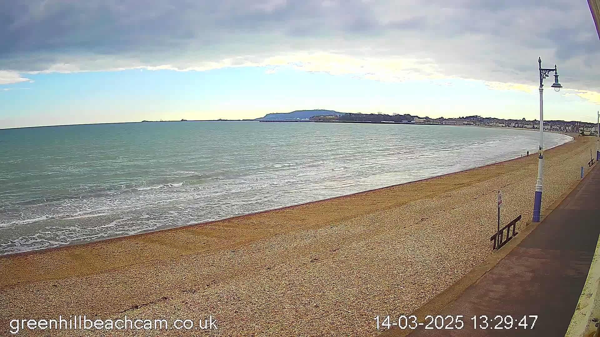 A sandy beach with gentle waves under a partly cloudy sky stretches along a calm sea to a distant coastline.