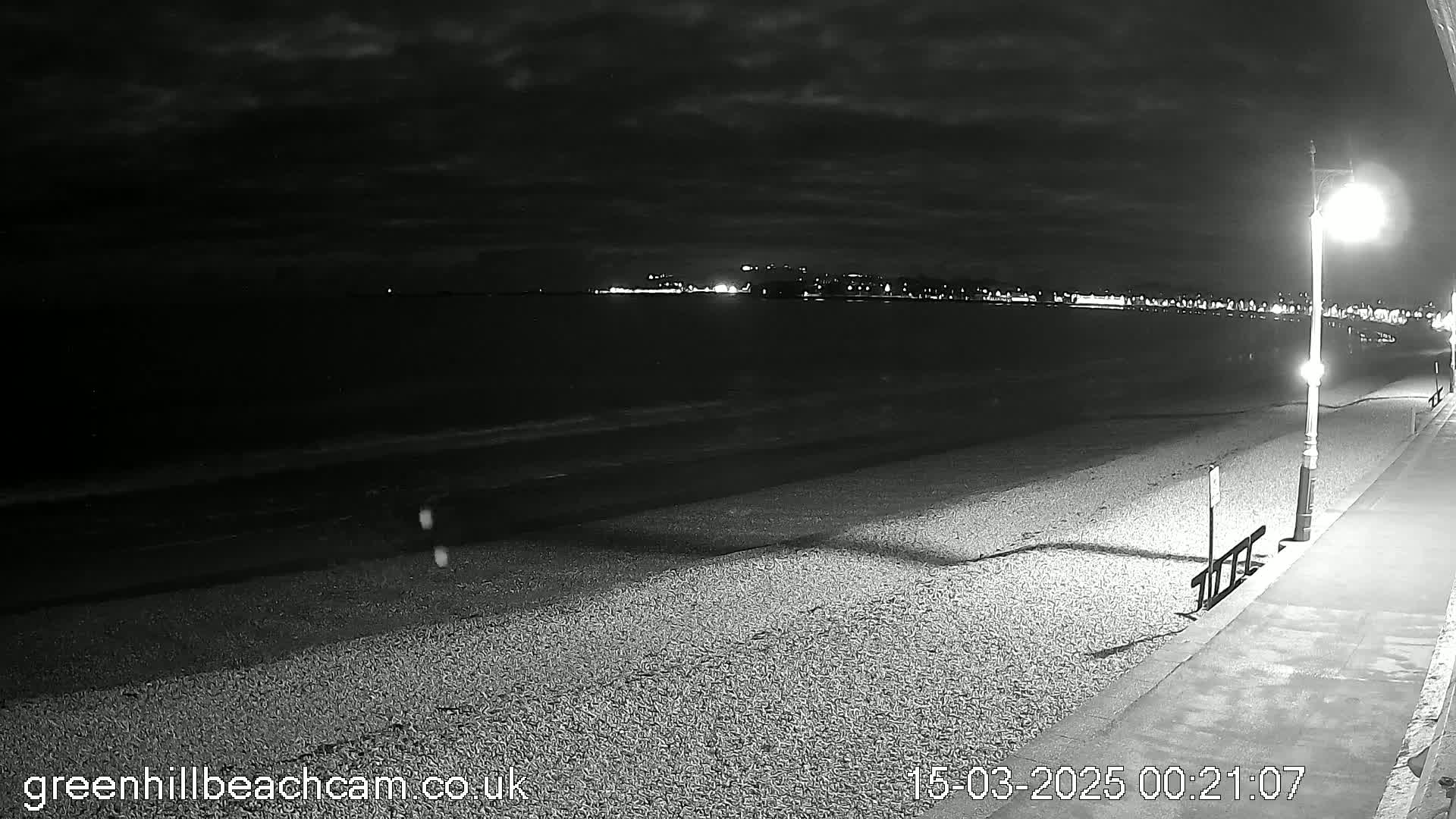 A nighttime view of a pebble beach and promenade, with a city illuminated in the distance under a cloudy sky.
