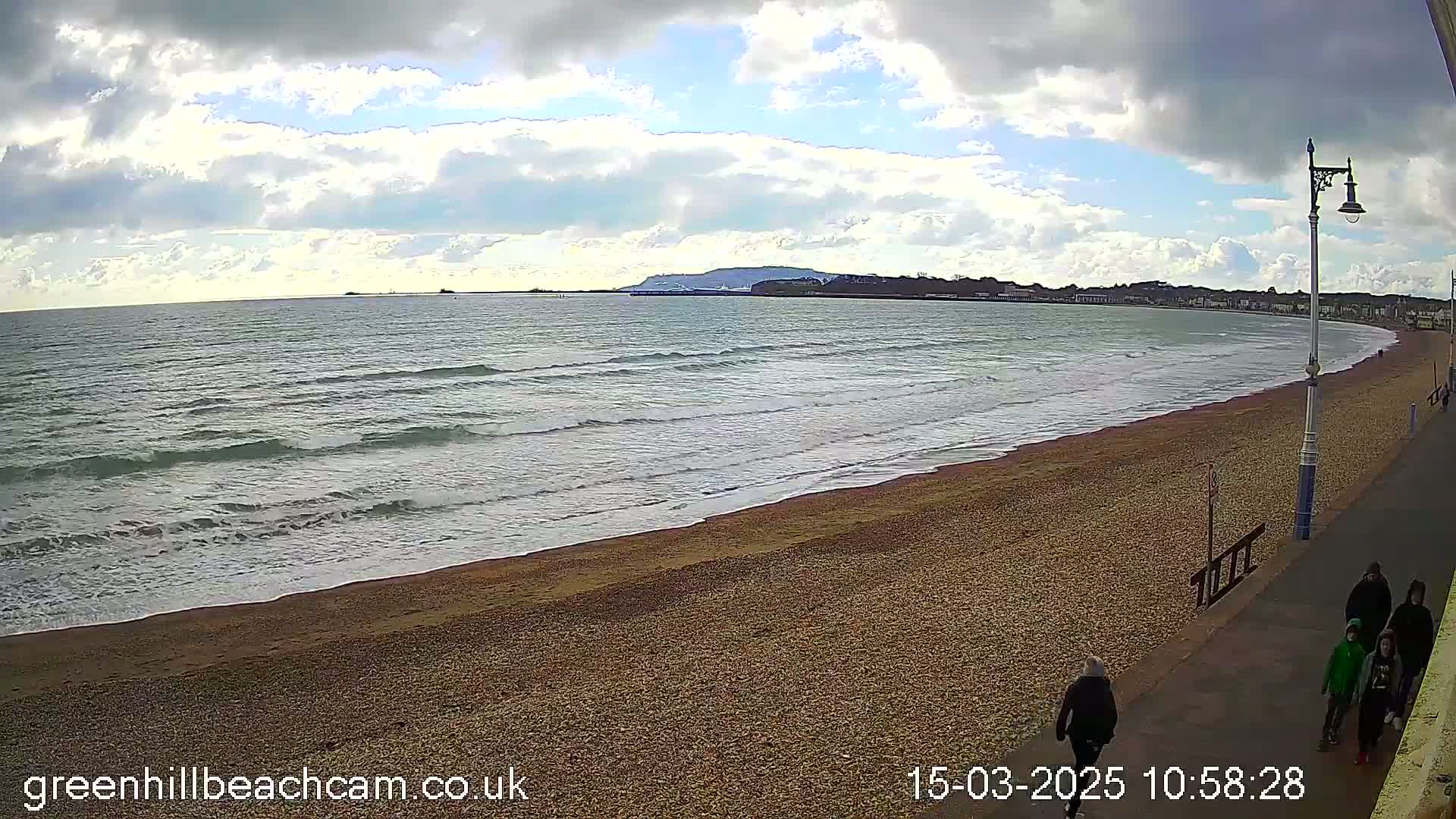 A pebbled beach with gentle waves under a partly cloudy sky, with a few people walking along a promenade.