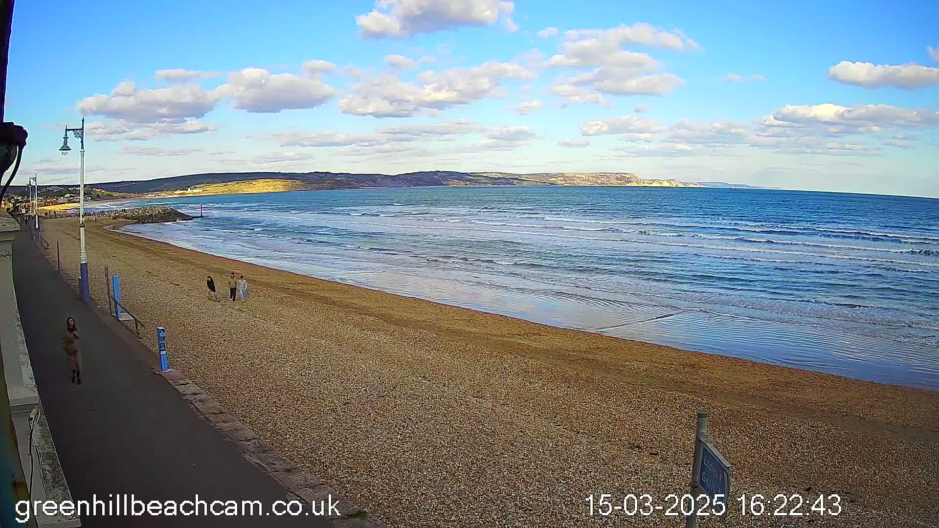 A sandy beach with a few people walking on it, under a partly cloudy sky, with waves rolling into the shore.