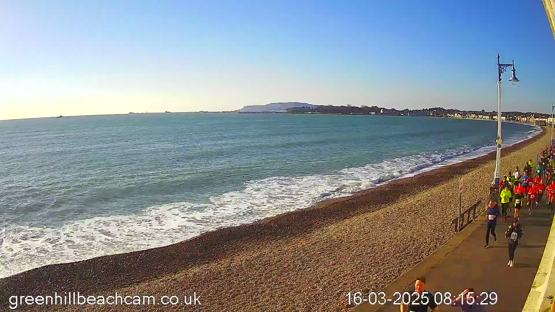 A group of runners is on a pebbled beach next to a calm sea under a clear blue sky.