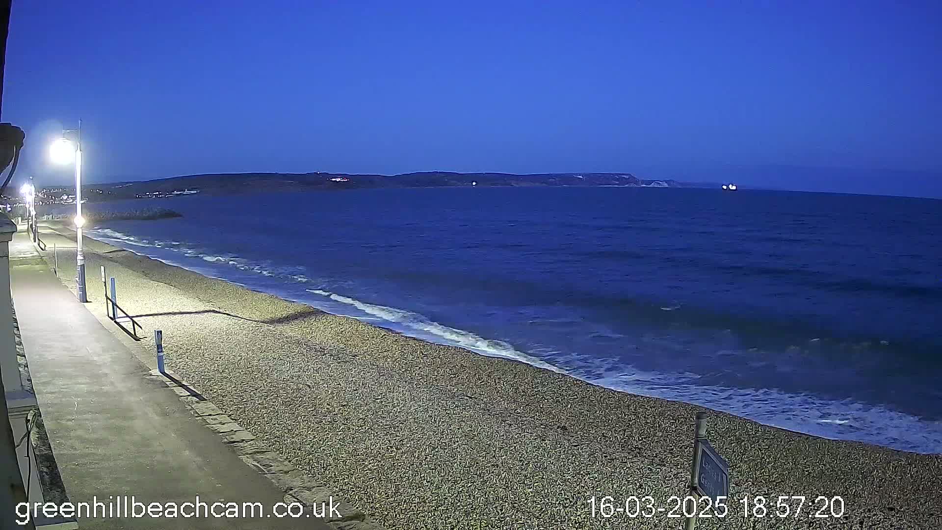 A pebble beach at twilight shows a calm sea with small waves lapping the shore, under a clear, dark blue sky.