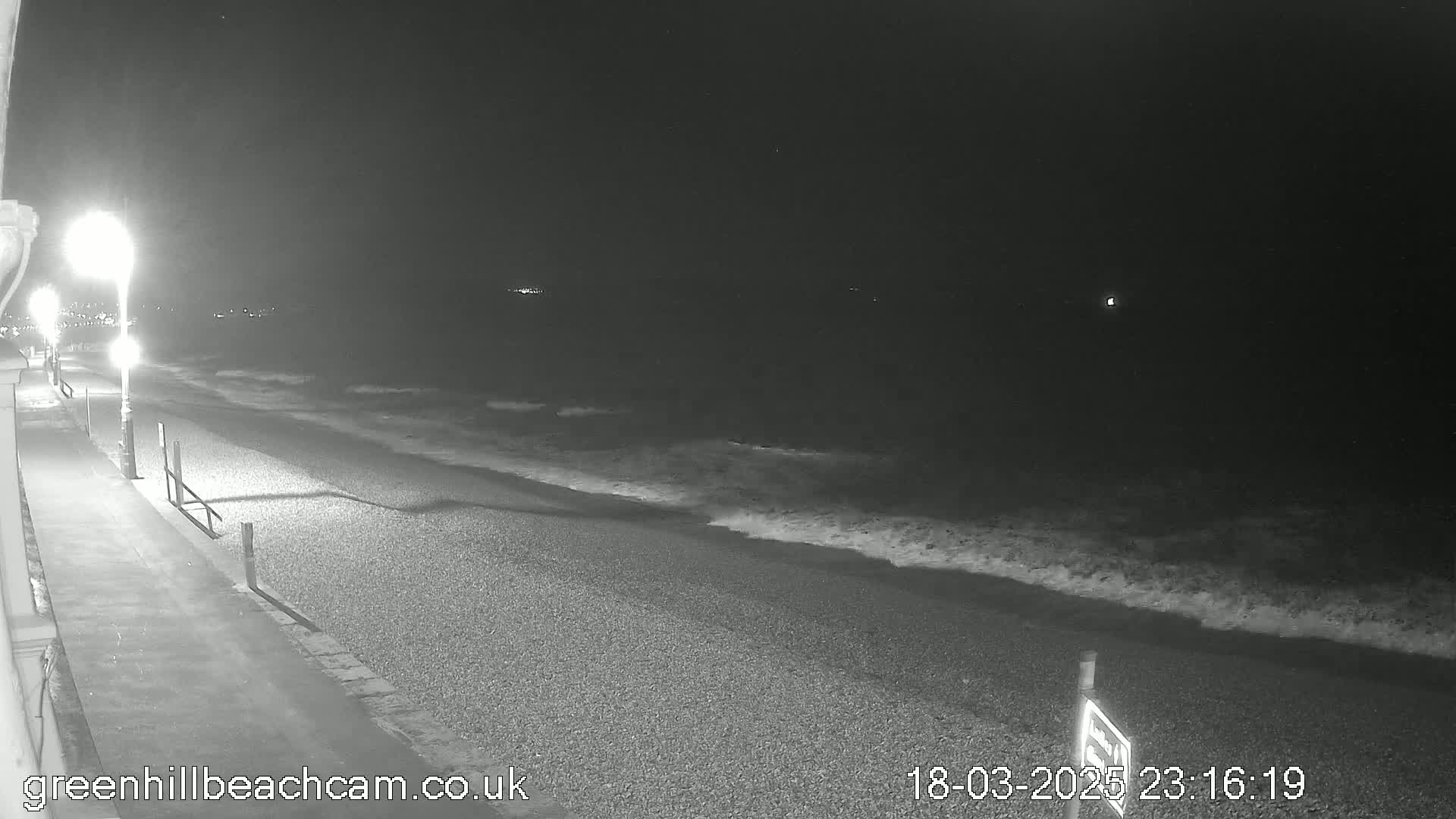 A dark, nighttime beach scene shows gently lapping waves on a pebbled shore, illuminated by streetlights along a paved walkway.