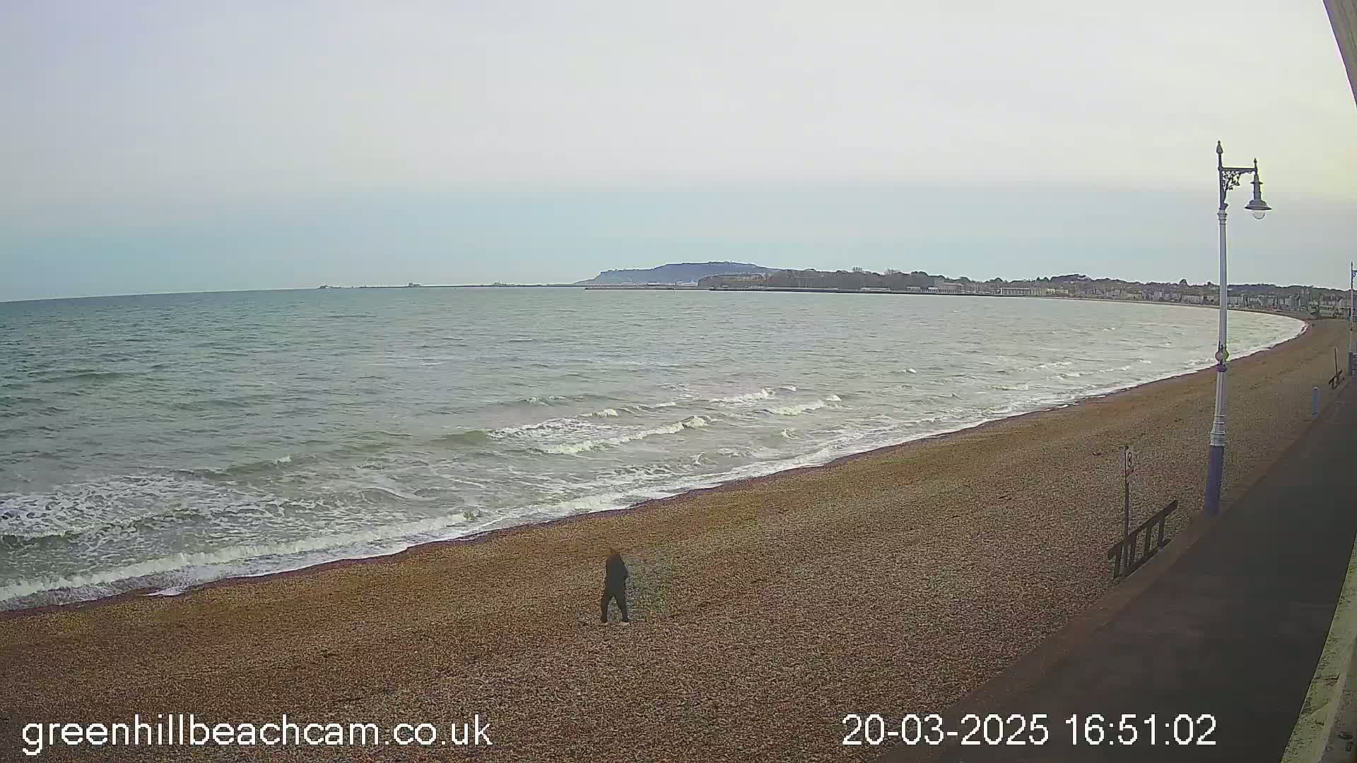 A person walks along a pebble beach with moderate waves and a calm, overcast sky in the background, where a town is visible in the distance.