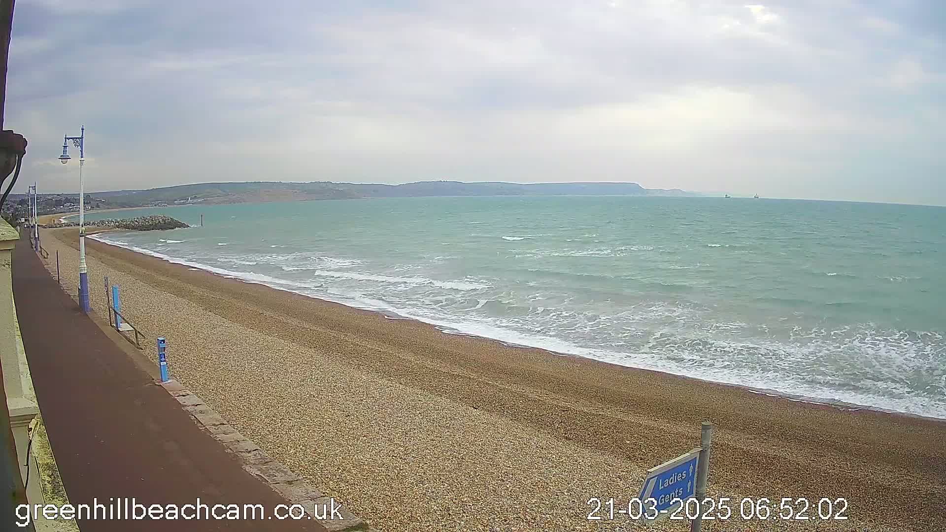 A pebble beach stretches along a calm sea under an overcast sky, with a paved walkway and distant hills visible.